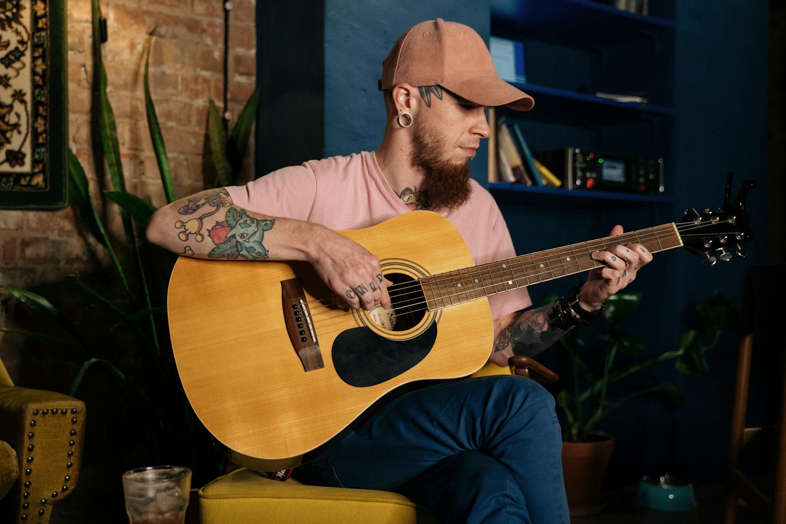 Bearded musician with tattoos playing acoustic guitar indoors. Captures artistic expression with a rustic interior.